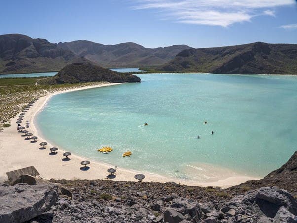 Playa con agua cristalina, palapas a su alrededor y montañas en el horizonte.