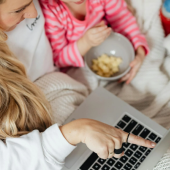 Madre e hija disfrutan de un momento de entretenimiento frente a una laptop. La madre señala la pantalla mientras la niña observa con interés. Ambas comparten una experiencia agradable y divertida.